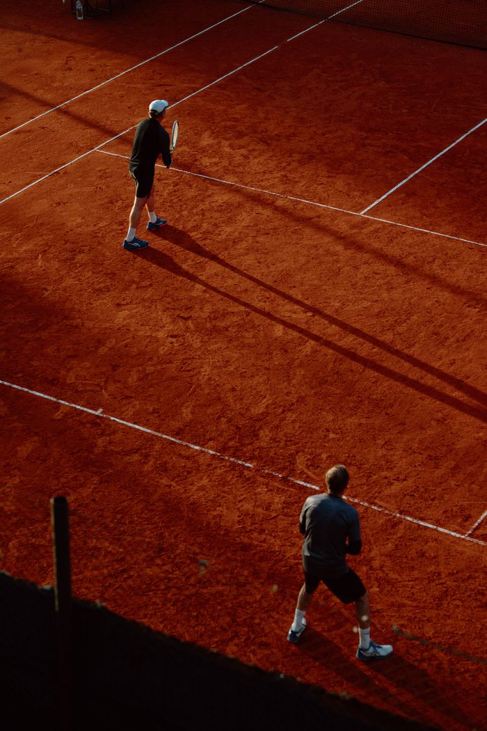 Home Two adults playing tennis on a red clay court during daytime. Shot from above, highlighting shadows.