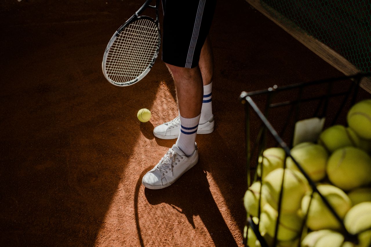 Contact A tennis player stands on a clay court, showing sneakers, racket, and tennis balls in sunlight.